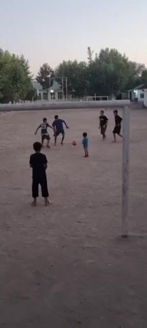 A group of boys are playing soccer on a dirt field in front of a goal. The sky is overcast and it appears to be late afternoon. The field is somewhat uneven and dusty. There are buildings and trees in the background. One boy is kicking the ball towards the goal.