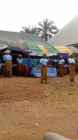 The video shows a group of African women performing music outdoors. The women are wearing matching outfits consisting of light blue shirts and long, patterned skirts. They also have yellow headwraps. Some of the women are playing drums, while one is singing into a microphone. There is a table with a sign that reads "ST ANTHONY OF PADUA IND/IE/ANU STATION". The ground is covered in what appears to be small, reddish-brown pebbles. There is a tent set up behind the group, and the overall setting appears to be a casual, outdoor gathering. Some blue plastic chairs are stacked at the edge of the scene. A palm tree can be seen in the background, and a building is partially visible behind the tent.