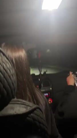 The video begins with a shot of the interior of a car, focusing on the back of a person's head with long brown hair. The interior light is on, casting a bright glow. The camera then pans to the right, revealing a young woman in the passenger seat looking at the camera. She is wearing a black hoodie and holding a pink phone. The camera then moves closer to her face, and she smiles briefly. The video returns to a shot of the car's interior, with the person in the driver's seat now visible. The dashboard and controls are illuminated, and the interior light is still on. The video ends with a blurred shot of the interior.
