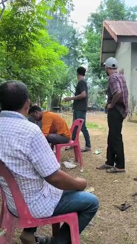 A group of men are gathered outdoors. In the foreground, a man wearing a plaid shirt and jeans sits on a pink plastic chair, his back to the camera. To his right, another man in an orange shirt is seated on a chair, with his head down. Further back and to the right, a man in a black shirt and jeans stands, holding something in his hand. In the middle ground, a man in a black shirt and a hat is leaning over a table covered with a red and blue cloth, on which a stack of white plates is visible. Behind him, another man in a brown shirt is crouched down. Several trees with white bark are prominent in the scene, with lush green foliage in the background. There is a small, rustic building with a corrugated roof on the left side of the frame. Cardboard boxes and some debris are scattered on the ground. The overall environment appears to be rural or semi-rural.