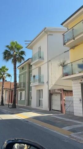 The video shows a view from inside a vehicle driving along a street lined with palm trees and buildings. The buildings are a mix of residential and possibly commercial, with varying colors and architectural styles. Some buildings have balconies with glass railings. The street is paved, and there is a sidewalk with a patterned border. The sky is clear and blue. The scene appears to be in a warm climate. There are no people visible in the video.