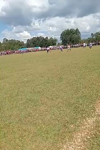 The video captures a soccer game in progress on a grassy field.  The sky is blue with scattered clouds. A crowd of spectators lines the edge of the field. Several players are running across the field, and the ball is in play. The camera is held by someone standing on the sidelines, who is partially visible in the frame. Their shadow is cast onto the grass.