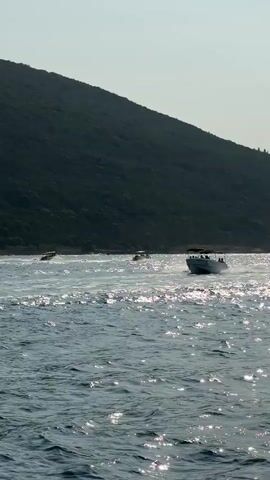 The video shows a body of water with several boats. In the foreground, the water is choppy and reflects the sunlight. In the background, a green, tree-covered hill dominates the scene. Several boats are visible on the water. One motorboat in the middle ground is kicking up a spray as it moves. Further in the background, other boats, possibly including a sailboat, can be seen. The sky is clear and the lighting suggests it is daytime.