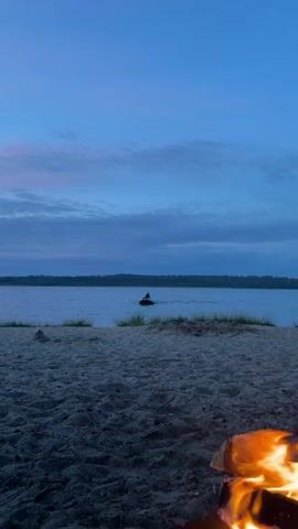A person is on a jet ski in the water. The water is calm with some grass growing near the shore. The shore is made of sand. There is a campfire in the foreground of the image. The sky is a light blue with some pink and white clouds. The background shows a treeline.