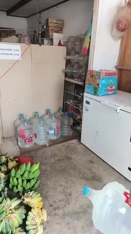The video shows a person carrying multiple large bottles of water. The person is wearing a t-shirt and shorts, and is walking from right to left in the frame. There are many bunches of bananas in the foreground. In the background, there are shelves stocked with various items, including more water bottles. The scene appears to be an indoor market or shop. The person is carrying two bottles of water at a time, one in each hand, and is seen placing them down near a collection of other water bottles. The person walks out of the frame and then returns to pick up more bottles. The floor is concrete, and there are some wooden structures in the room. The lighting is bright, suggesting daytime.