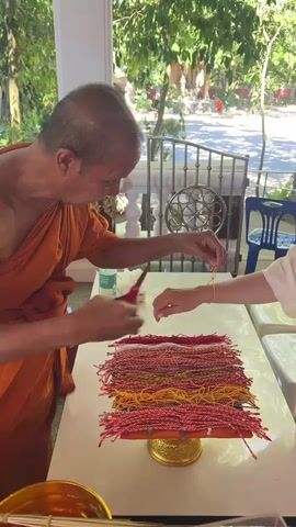 A Buddhist monk in an orange robe is performing a ritual with a woman in a light-colored outfit, likely in Baan Hua Thanon, Thailand. The monk is tying a red and white string bracelet around the woman's wrist. In front of them, on a white table, is a large pile of similar bracelets, neatly stacked on a golden pedestal. The scene appears to be outdoors, with lush green trees and foliage visible in the background, suggesting a warm, sunny day. The monk has a gentle expression, and the woman seems engaged in the ritual. The setting looks like a temple or a similar spiritual site, with decorative pillars and a metal fence visible. The activity is a common Buddhist practice, where blessed threads are tied to offer protection and good fortune.