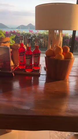 The video pans across a table setting with three bottles of Aperol, a bowl of oranges, and a lamp. In the background, there is a view of islands and the sea, with a fence adorned with pink and white flowers. The scene appears to be set at sunset or sunrise, with soft lighting.