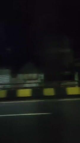 The video shows a night-time view from a moving vehicle, possibly a car. The camera is facing to the side, capturing the blur of buildings and streetlights as they pass. The scene is quite dark, illuminated primarily by the artificial light sources and the headlights of other vehicles. There is a yellow and black barrier or wall alongside the road. Due to the motion and darkness, details of the buildings and objects are difficult to discern clearly.