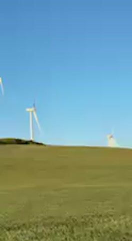 The video captures a serene landscape featuring a wind farm on a sunny day. Several large wind turbines with white blades stand tall on a green, rolling hillside, extending into the distance. The sky is a clear, vibrant blue, indicating fair weather. The sunlight illuminates the turbines, giving them a slight golden glow. The camera moves gently, panning across the scene, showcasing the scale of the wind turbines against the expansive blue sky and green hills. The blades of the turbines are visibly rotating, driven by the wind, which is also audible throughout the clip. In the background, other wind turbines can be seen dotting the distant hills, appearing smaller due to perspective. There are no people or visible text in the video. The setting appears peaceful and natural, highlighting renewable energy infrastructure.