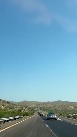 A long, straight highway stretches into the distance under a vast, clear blue sky. The road is asphalt, with a yellow line marking the left edge and a white line indicating the right lane. Several cars are visible on the road, traveling in the same direction as the viewer. The landscape on either side of the highway is arid and mountainous, with sparse desert vegetation such as scrub brush and cacti. The sun appears to be high in the sky, casting a bright light on the scene. There is a green highway sign with white text visible on the right side of the road. The overall impression is one of a long, open-road journey through a desert environment.
