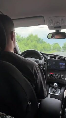 The video shows the interior of a car from the perspective of a passenger in the back seat. A man wearing glasses and a dark t-shirt is driving. He is looking straight ahead. The car is on a road surrounded by trees. The dashboard of the car is visible, including the steering wheel, a central infotainment screen displaying a song title and album art, and climate controls. The gear shift is also visible. The view through the windshield shows the road ahead and other cars in the distance. The sky is overcast.