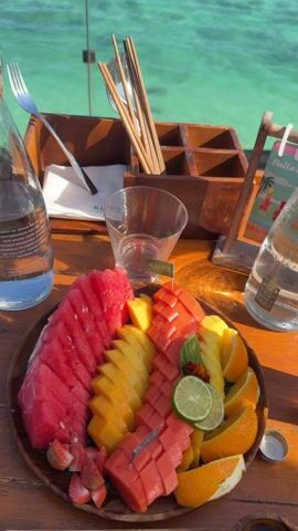 The video shows a plate of tropical fruits on a wooden table, along with a bottle of water, a glass, a silverware holder, and a menu. The fruit plate contains watermelon, mango, papaya, orange slices, strawberries, and a lime wedge. The backdrop is a scenic view of a turquoise ocean and a clear blue sky. A person is seen wading in the shallow water near the shore. The scene suggests a leisurely meal or snack enjoyed in a coastal setting.