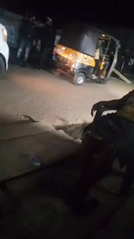 The video shows a nighttime scene with a white van parked on a dirt road. The van's side door is open, revealing some people inside. In the background, there are other people standing near a three-wheeled vehicle. The person filming is sitting, with their arm visible in the foreground. The overall lighting is dim, making it difficult to see details clearly.