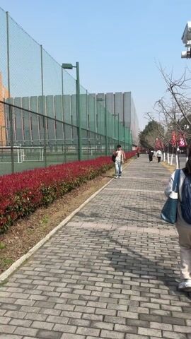 The video shows a sidewalk paved with grey bricks, with red bushes lining the left side, bordering a green sports field enclosed by a high fence. People are walking along the sidewalk in both directions. A man wearing a mask and carrying a bag walks away from the camera, and a woman with a blue backpack walks towards the camera on the right side of the frame. In the background, there is a large, modern building and trees lining the sidewalk on the right. The sky is clear and blue.