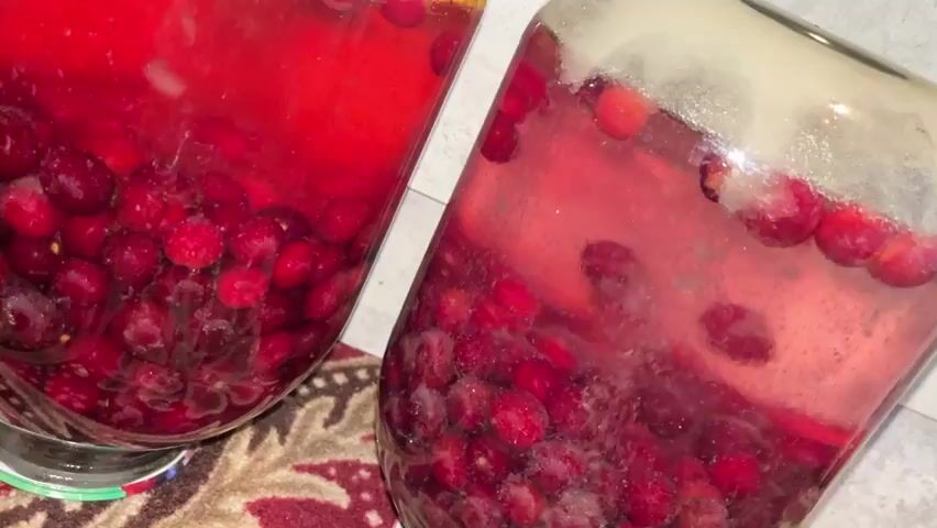 The media file shows a close-up, still-life view of three large glass jars filled with a vibrant red liquid and numerous small, dark red berries. The berries are concentrated at the bottom of each jar, with the liquid filling the upper portion. Some berries are floating near the surface. The jars are placed on a patterned rug with red and cream colors. The lighting is somewhat dim, suggesting an indoor setting. The focus is on the jars and their contents, creating a sense of preserved fruit or a beverage being made. There is no visible text. The scene is set in Bujum, Kyrgyzstan. The overall impression is one of homemade preservation or a traditional beverage preparation.