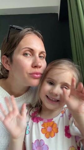 A woman and a little girl wave hello into the camera. The woman wears a white sweater and has her hair pulled back; her sunglasses are perched on her head. The little girl has light hair and wears a floral dress. They are indoors, and behind them is a dark green wall and a green curtain. The woman kisses the girl on the head.