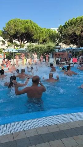 The video shows a group of people in a swimming pool, some in the water and some standing on the edge. The people in the water are splashing and moving around, seemingly participating in an activity like water aerobics. There are several adults and children present. The environment appears to be a resort or a public pool area, with lounge chairs, umbrellas, and lush green trees visible in the background. The water is blue and clear, with visible splashes and ripples. The lighting suggests it is daytime, with clear skies. Some individuals are wearing swimwear. There is a sense of active participation and enjoyment among the group.