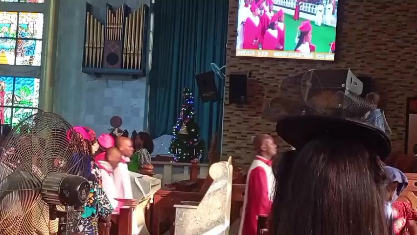 The interior of a church is shown with several people present. The stained-glass windows on the left wall let in light. An organ is installed on the wall above, next to a long green curtain. A small Christmas tree sits on the floor. A large television screen on the right wall shows images of men in robes. People are standing in the foreground, some wearing colorful hats. Young people in white robes stand near wooden benches. A large fan is running. A woman with dark hair and a large black hat stands in front of the camera.