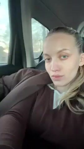 A woman sits in the back seat of a car, looking at the camera. She has fair skin, blonde hair pulled back in a ponytail, and green eyes. She is wearing a brown sweater and a white undershirt, with a brown jacket draped over her shoulders. Outside the car window, a bright sunset illuminates a street. The car is moving, and the scene outside the window changes. The woman maintains a calm expression, slightly tilting her head. The car's interior is visible, with black leather seats and gray panels.