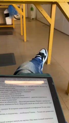 A close-up shot shows a person's legs and a tablet computer with an open book on the screen. The person is wearing blue jeans and white sneakers with black accents. The tablet is resting on the person's lap, and their feet are crossed. The background is out of focus but appears to be an indoor or covered outdoor space with yellow furniture and some shelving. The scene is brightly lit.