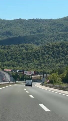 A white van travels down a multi-lane highway. On either side of the highway, there are green, tree-covered hills. Overhead, a blue sky is visible. The highway has white lane markers and a guardrail on the right side. There are several road signs visible, including speed limit signs and signs with red and white chevron patterns. The van is moving away from the camera.