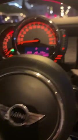The video begins with a close-up shot of a car's dashboard, specifically a speedometer with red illuminated markings and a digital display showing time and mileage. The steering wheel, bearing the "MINI" logo, is also visible in the foreground. The camera then pulls back slightly, revealing the view through the windshield at night. The car appears to be in traffic, with other vehicles visible ahead and to the sides. The focus shifts to a bright red and blue sports car in front, its taillights illuminated. The surrounding environment shows streetlights and buildings, indicating an urban setting at night. The video continues with a series of shots that maintain this perspective, showing the traffic and the sports car ahead as the vehicle moves forward slowly. The dashboard lights and the street scene are consistently visible.