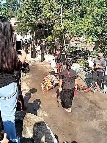 The video shows an outdoor scene with several people. In the foreground, a person with long dark hair is filming with a smartphone. They are wearing a black t-shirt and light blue jeans. To their right, another person is dressed in black, holding a broom. In the background, a group of people are gathered, some in black clothing. There are trees and foliage surrounding the area. In the middle ground, there are individuals wearing traditional costumes, including one person lying on the ground with another standing over them. Further back, there are more people dressed in black, some standing and some seated. A portable stage or setup covered with a blue tarp is visible on the right side of the frame, with audio equipment, including speakers and a purple amplifier, on display. The ground appears to be a dirt path. The lighting suggests it is daytime.