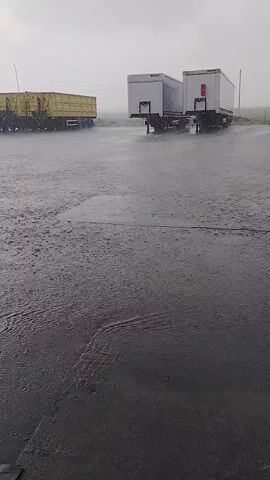 This video captures a wide shot of a large outdoor area, likely a truck stop or industrial yard, during a heavy downpour in Karapelit, Bulgaria. The ground is entirely covered in water, with visible puddles and ripples from the intense rain.

Various trucks and trailers are parked across the scene. In the foreground, the ground is dark and glistening with water, reflecting the overcast sky. In the middle ground, numerous commercial vehicles are visible, including white semi-trucks, some pulling white box trailers, others with yellow dump trailers, and at least one with a tank trailer. Several white vans are also parked among the larger vehicles. In the background, there are utility poles and what appears to be part of an industrial structure or power lines.

The rain is the dominant element, creating a sense of a washout. No people are visible. A partial sign with the word "EAT" is faintly discernible on the left side of the screen at 0:03, suggesting a rest area or food establishment nearby. A wooden crate or barrier is present in the right foreground of the last frame.