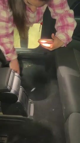 A woman wearing a pink and white plaid jacket and grey pants is vacuuming the inside of a car. She is using a handheld vacuum cleaner in her left hand and a cell phone for light in her right. The car’s interior is black and grey. The vacuum cleaner is also black. The woman is bent over, focusing on cleaning the floor area in the back seat. She has long, dark hair. The lighting is dim, with the cell phone providing the primary light source.