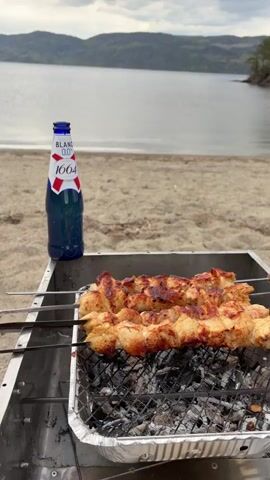 The video shows a beach barbecue scene. Two skewers of grilled chicken sit on a grill over glowing coals in a foil pan. A bottle of Blanc 1664 beer stands beside the grill. The beach is sandy, and a calm lake stretches out to distant, rolling hills under an overcast sky.