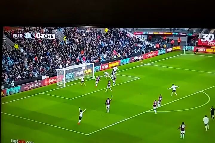 The video shows a close-up of a television screen displaying a soccer game. The score is Burnley 1, Chelsea 1. A player wearing a white jersey with the number 7 on the back is walking towards the camera, then turns to celebrate with another player. The other player is wearing a white jersey with the number 8. They both are yelling and pointing. The background includes the green soccer field and a red and yellow advertising board. A logo that says "30 Years" is on the upper right corner of the screen.