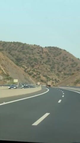 The video shows a view from inside a vehicle traveling on a highway. The highway has multiple lanes, and is divided by concrete barriers and white dashed lines. The road curves to the right and is flanked by a steep, rocky hillside covered in sparse green vegetation. There are a few vehicles visible on the highway, including a dark-colored car on the left side of the road and a truck in the distance on the right side of the road. The sky is clear and pale blue.