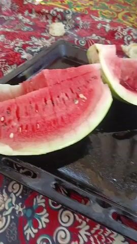 A close-up shot shows two slices of watermelon on a black baking sheet. The watermelon is bright red with black seeds. The baking sheet is resting on a patterned fabric. In the background, a person wearing blue pants is partially visible.