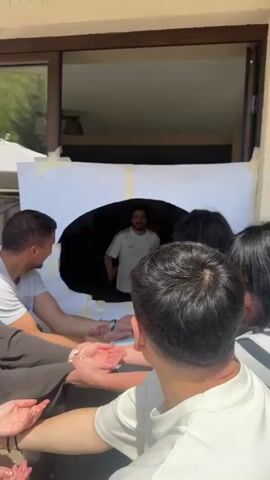 A group of five young adults, including both men and women, are gathered outside near a building with a large window. They are participating in a game or activity where one person is reaching their arms through a hole in a large white sheet, while the others, standing close together, are touching the outstretched hands. The person behind the sheet is quickly pulled forward, causing the group to react with surprise and laughter. The environment includes natural light, suggesting an outdoor setting. The clothing worn by the participants is casual. The mood appears lighthearted and fun.