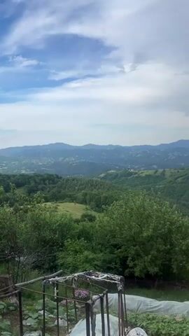 The video pans across a lush green landscape of rolling hills and mountains under a partly cloudy sky. In the foreground, there is a makeshift greenhouse or cold frame structure with white plastic sheeting and metal poles. Some plants are visible growing around it. The camera moves slowly, revealing more of the vast, verdant scenery with a sense of distance and depth. The overall impression is one of rural tranquility and natural beauty.