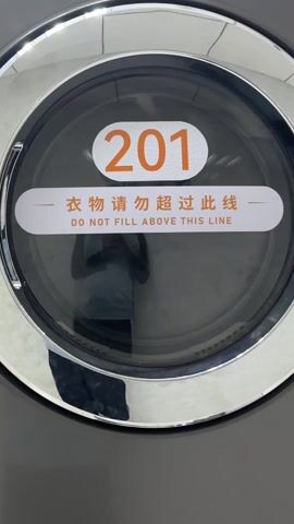 A close-up shot focuses on the door of a commercial-grade washing machine or dryer. The machine is metallic gray and features a large, round door with a chrome-colored trim. On the door, there's a sticker with the number "201" in bright orange, placed above a horizontal line. Below this line is a white rectangle with black text in both Chinese and English, which reads "衣物请勿超过此线" and "DO NOT FILL ABOVE THIS LINE." The glass of the door is reflective, showing a faint image of a person holding a phone, likely taking the video or picture. The lighting is bright and uniform.