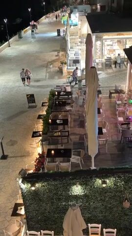 A night view of a seaside promenade with a restaurant. The promenade is paved with cobblestones and lined with lampposts. There are several people walking on the promenade. To the right, a restaurant with outdoor seating is visible. The tables are set with chairs and umbrellas. The scene is illuminated by artificial lights.