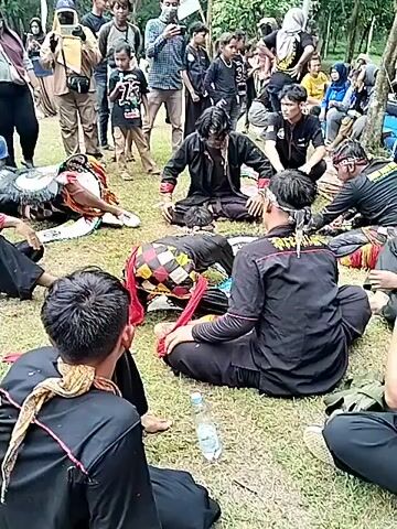 The scene takes place outdoors on a grassy area, with trees visible in the background. A group of people, mostly young men, are gathered. They are dressed in black attire with red accents. Some are seated on the ground in a circle, while others stand around observing. Several individuals are lying face down on the grass in the center of the circle. People are recording the scene with cameras and phones. There are water bottles scattered around the seated individuals. A mix of adults and children are watching from the periphery. The atmosphere seems to be a combination of curiosity and concern.