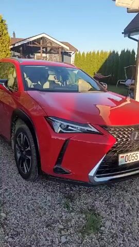 The video shows three vehicles parked on a gravel driveway in front of a house. The first vehicle is a red Lexus UX crossover. The second vehicle is a gray Volkswagen Touran wagon with a roof box. The third vehicle is a red, yellow, and black Can-Am Maverick X3 side-by-side. The house is in the background, surrounded by trees and bushes. The weather appears sunny.