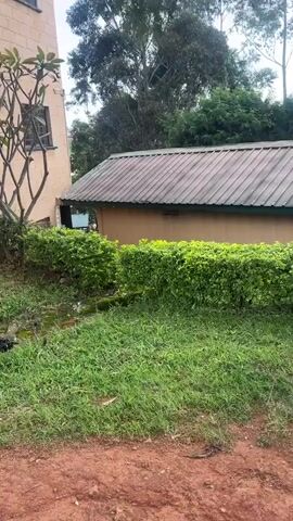 This video shows an outdoor view of a residential or institutional complex in Gembu, Nigeria. The scene features a multi-story, light peach-colored building with numerous windows, some of which have protective bars. In front of the building, there are manicured green hedges and a leafless tree. A smaller, single-story building with a brown, corrugated metal roof is also visible.

The ground is covered with a mixture of grass and reddish-brown dirt, indicating an unpaved area. In one section, there's a pile of black trash. A signpost with some faded text is planted near the building. Further into the complex, other similar buildings can be seen, along with parked cars and trees in the background. The sky is partly cloudy, suggesting daytime.

The video pans across the area, showing different angles of the buildings and their surroundings. In the final segment, a man wearing a green t-shirt and a black cap is seen standing next to a motorcycle. He appears to be on a dirt path in front of one of the buildings. The overall impression is one of a functional, somewhat rural or semi-urban environment. The presence of the motorcycle and the unpaved roads suggest a locale where such elements are common.