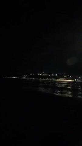 A very dark video shows a distant coastline illuminated by numerous small lights. The lights appear to be from buildings or streetlights along the coast, which extends from the left side of the frame towards the right. The sea is visible in the foreground, reflecting some of the distant lights, creating a shimmering effect. The overall impression is of a town or city by the sea at night. The video appears to be shaky and out of focus in some areas, suggesting it was filmed while moving.