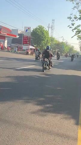 A group of motorcyclists ride down a paved road. In the background, a Pertamina gas station with red and white signage stands to the left. Trees and utility poles line the right side of the road. The sky is clear and blue. The motorcyclists are all wearing helmets. The road is in good condition. The sun is shining, casting shadows on the road.