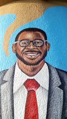 The video shows a mural on a cylindrical surface. The mural depicts a smiling man with dark skin, wearing glasses, a grey suit, a white shirt, and a red tie. He is standing with his hands in his pockets. The background consists of abstract shapes in various colors including blue, yellow, pink, and green. The cylindrical surface itself is a light brown color above the mural.
