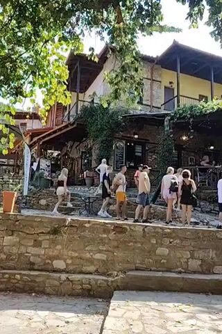 A group of people are walking up stone steps towards an outdoor cafe. The cafe has a building in the background with a yellow upper story and stone lower story. There are tables and chairs set up outside under a canopy of trees and vines. Some people are seated at the tables. The ground is paved with stones.