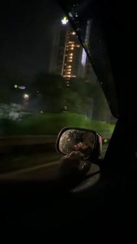 This is a first-person perspective video taken from inside a car at night. The camera is pointed out the passenger side window. In the side mirror, a person's arm and hand are visible, holding a phone. The phone screen reflects an image of the person's face, and they appear to be taking a selfie. The car is moving, indicated by the blurred scenery of a city skyline with tall buildings lit up at night, and greenery alongside the road. It seems to be raining, as there are water droplets on the car's window and the side mirror. The audio is of a person speaking, though the content is not fully clear in the provided snippet.