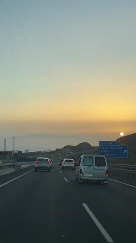 The video shows a view from inside a vehicle traveling on a highway at dusk. The sky is a gradient of pale blue and warm yellow, indicating the sun is setting or has recently set behind distant hills. Several cars are visible on the highway, traveling in the same direction. In the foreground, a silver van is in the right lane. To the left, a white sedan and other vehicles are visible in the lanes. A blue highway sign is visible on the right side of the road, with text including "1000 M" and place names like "Los Cristianos" and "El Teide". The environment appears to be a mountainous or hilly region with some sparse vegetation. The overall atmosphere is calm and transitional, typical of a journey during the evening hours.