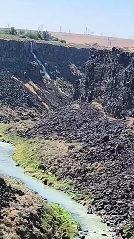 A video shows a river flowing through a canyon with a waterfall cascading down a rocky cliff. The canyon walls are covered in dark rocks, and vegetation grows along the riverbanks. In the background, wind turbines are visible on a distant ridge. The sky is clear and blue.