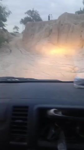 The video appears to be shot from inside a moving vehicle, driving on a rugged dirt road. The road is flanked by steep, eroded, earth banks. The lighting is dim, possibly dusk or dawn, and the vehicle's headlights are on. Two people can be seen standing on the top of one of the earth banks. The dashboard of the vehicle is visible, with some papers on top.