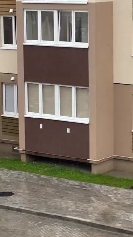 The video shows a section of an apartment building with a balcony. The building's facade is a mix of beige and dark brown. There are multiple windows visible, some of which are open. Rain is falling, as evidenced by the wet pavement and streaks on the building. A drainpipe is visible on the side of the building. A small patch of grass is at the base of the building. A car is partially visible in the bottom left corner.