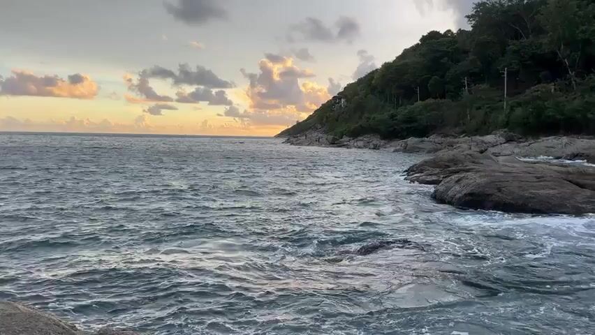 This video captures a serene coastal scene at sunset in Rawai, Thailand. The primary subject is the ocean, with waves gently crashing against a rocky shoreline. Lush green vegetation covers a hillside that slopes down towards the sea, suggesting a tropical or subtropical location. The sky is painted with soft hues of orange, yellow, and pink from the setting sun, with scattered clouds adding texture and depth. The water appears choppy in places, indicating a moderate breeze. The overall mood is peaceful and tranquil, evoking a sense of natural beauty and calm. No people or specific objects are clearly visible, beyond the natural elements of the landscape. The time of day is clearly sunset, with the warm, diffused light characteristic of that time. The presence of the ocean, tropical vegetation, and the warm sky confirm the location as Rawai, Thailand.